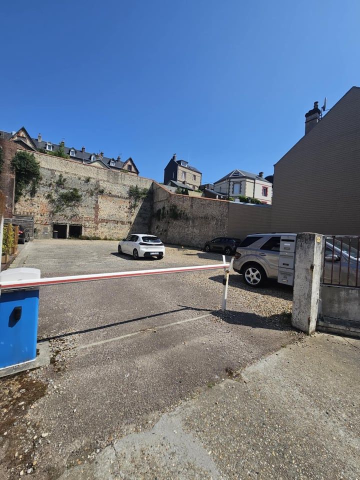 Parking avec voiture blanche, mur de pierre et poubelle bleue sous un ciel dégagé