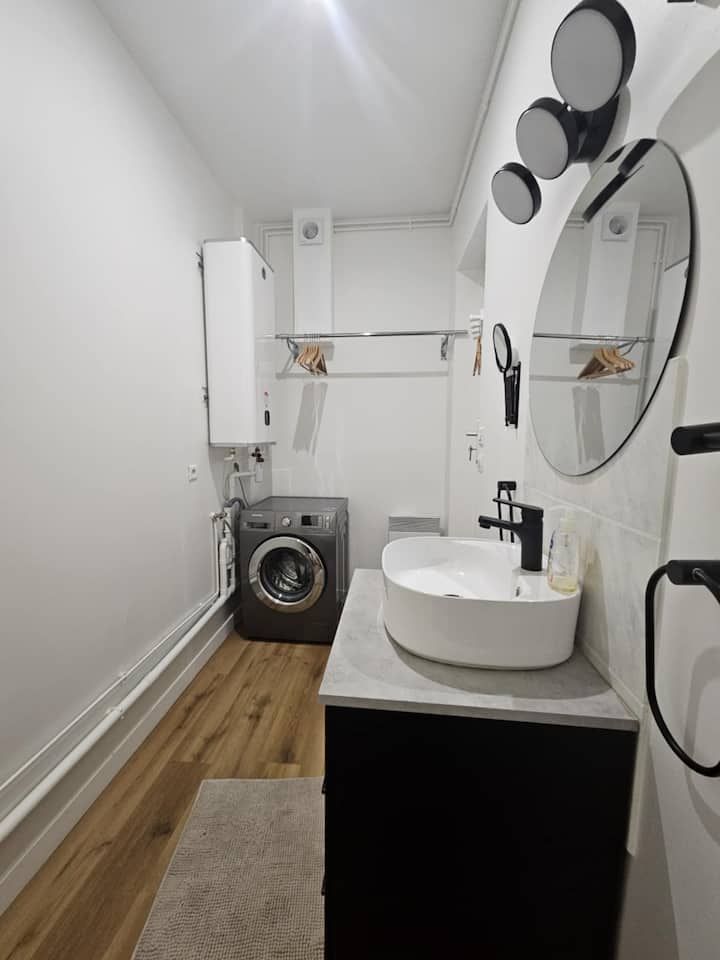 Buanderie étroite et blanche avec lave-linge, miroir rond, lavabo et tapis de sol en bois