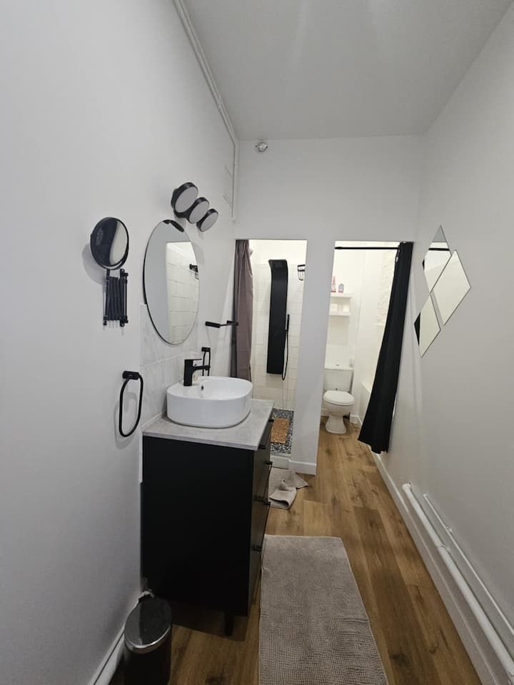 Couloir étroit et blanc avec parquet, miroir, lavabo et appliques murales menant à une porte.