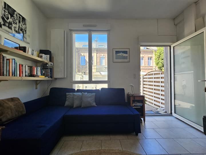 Petit salon avec canapé bleu, étagères, fenêtre et porte ouverte donnant sur un balcon.