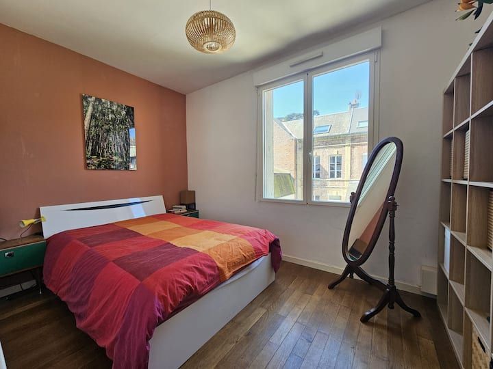 Chambre lumineuse avec un couvre-lit rouge-orangé, une lampe de fenêtre, un miroir et une étagère.