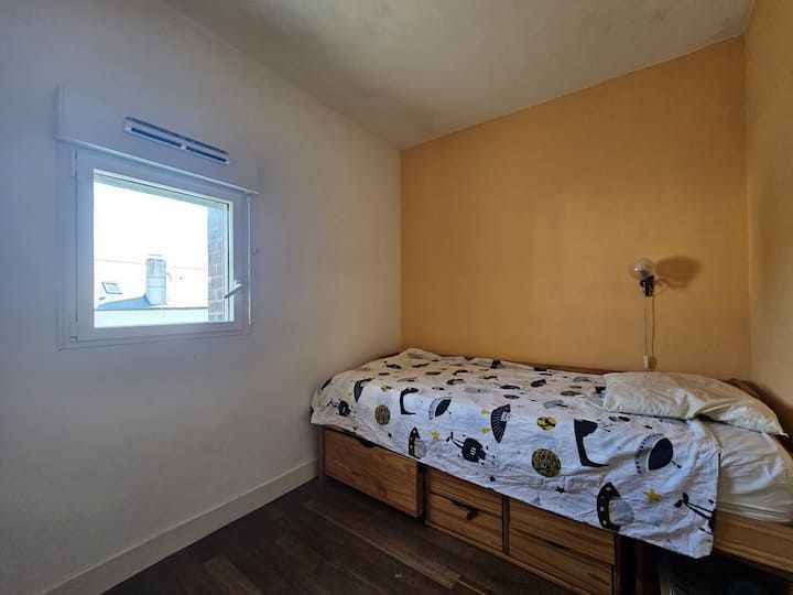 Petite chambre avec un lit simple, une fenêtre, des murs beiges et des tiroirs de rangement en bois en dessous.