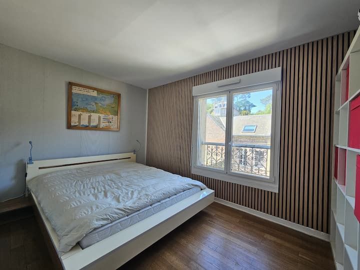 Chambre lumineuse avec lit blanc, papier peint à rayures et grande fenêtre avec rideaux.
