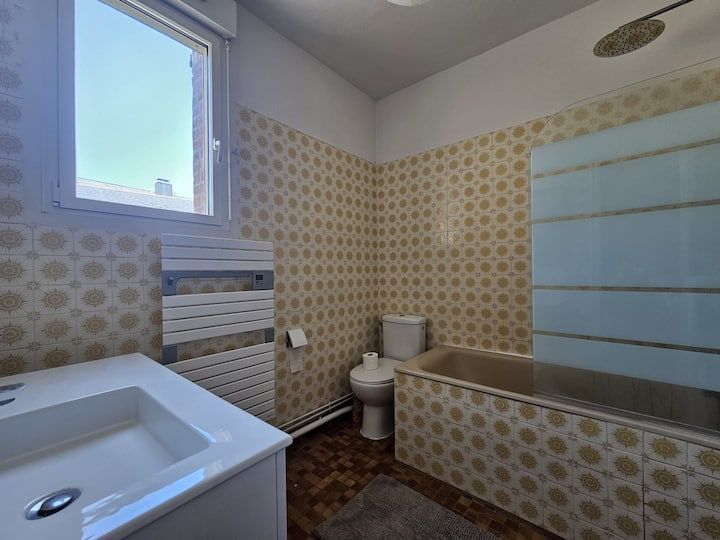 Petite salle de bains avec lavabo, toilettes, baignoire, fenêtre et murs carrelés de carreaux beiges à motifs.