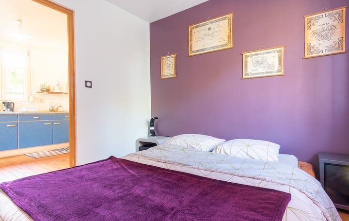 Chambre avec mur violet, lit, tableau encadré et porte donnant sur une cuisine avec des placards bleus.