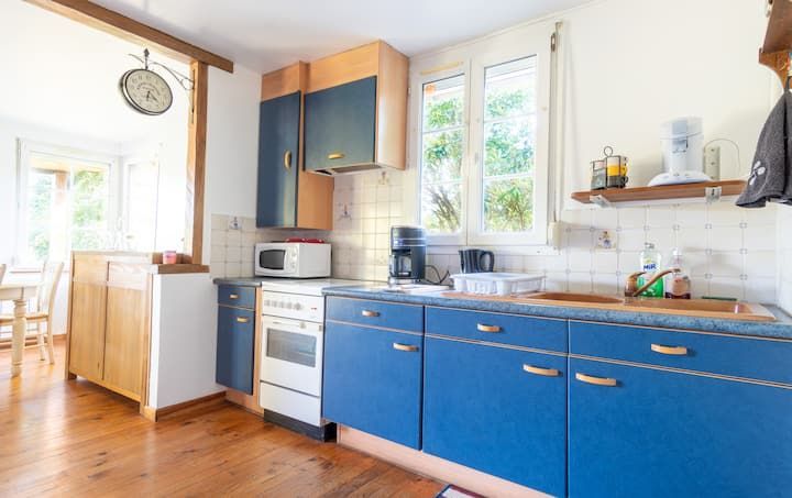 Cuisine avec placards bleus, électroménager blanc et fenêtre. Sol en bois et murs clairs.
