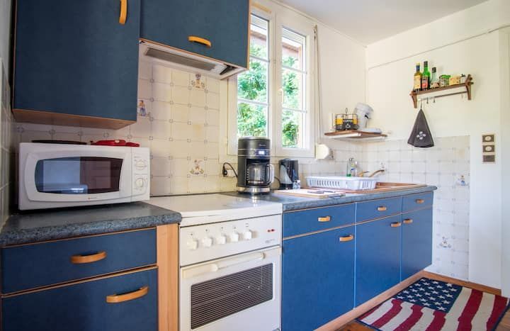 Petite cuisine avec placards bleus, électroménager blanc et fenêtre donnant sur la verdure. Tapis aux couleurs du drapeau américain.