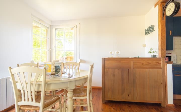 Salle à manger avec table ronde, quatre chaises et buffet en bois, éclairée par la lumière du soleil entrant par les fenêtres.