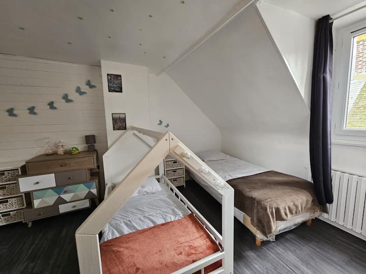 Chambre mansardée avec deux lits simples, murs blancs, commode en bois et fenêtre avec rideaux sombres.