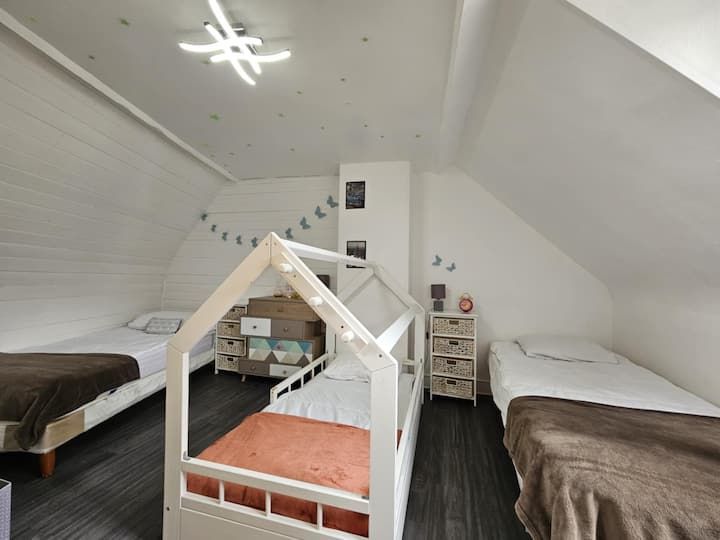 Chambre mansardée lumineuse avec trois lits, murs blancs en pente et parquet foncé