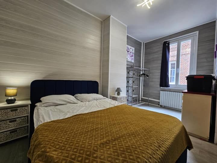 Chambre moderne avec lit défait, couverture moutarde, lampe de chevet et grandes fenêtres.