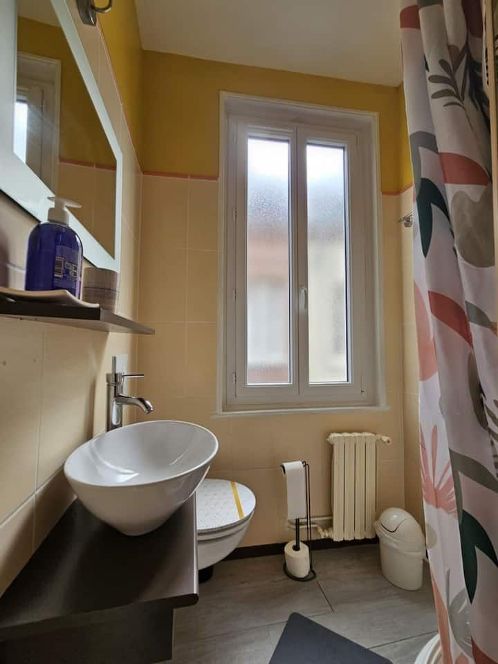 Petite salle de bains avec lavabo, toilettes, fenêtre, radiateur et rideau de douche à motifs floraux.