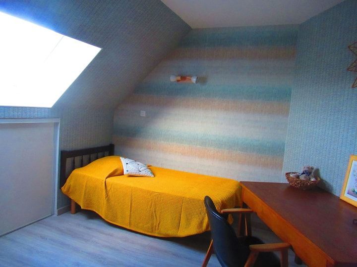 Chambre avec un lit, un bureau et un plafond mansardé. Couvre-lit jaune, mur à rayures bleues et oranges et porte blanche.