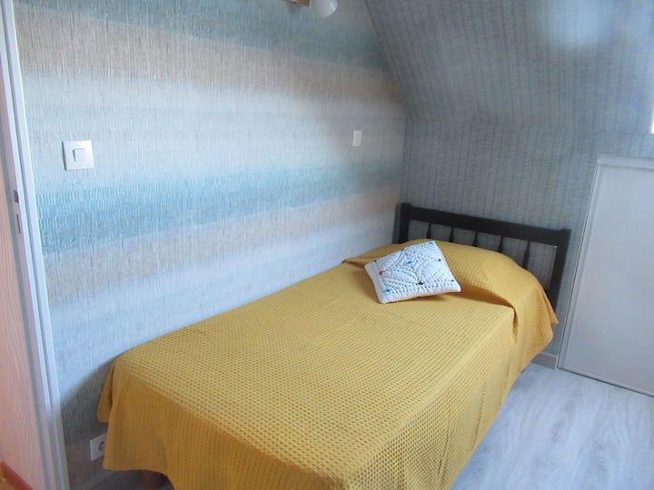 Chambre avec un lit simple, un couvre-lit jaune, un mur à motifs et un petit oreiller blanc.