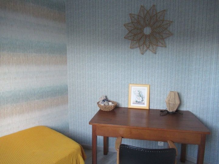 Coin cosy dans la chambre avec bureau, tableau et étoile tissée au mur. Couvre-lit jaune et papier peint à rayures bleu clair.