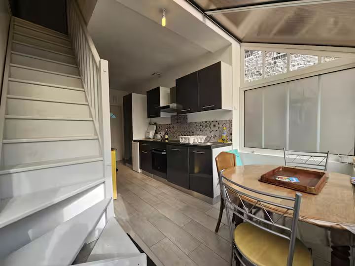 Petite cuisine et coin repas avec escalier, placards noirs et table près de grandes fenêtres.