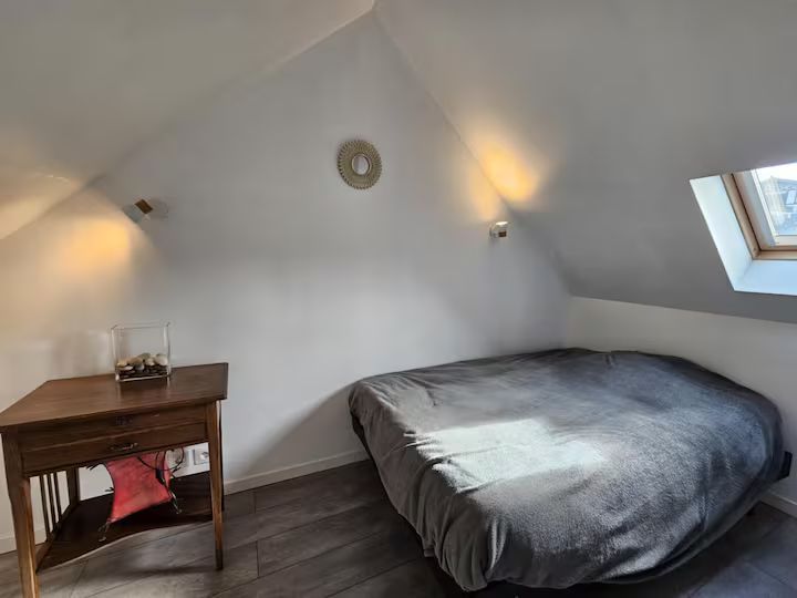 Petite chambre mansardée avec un lit, un bureau en bois et des murs blancs inclinés sous des fenêtres de toit.