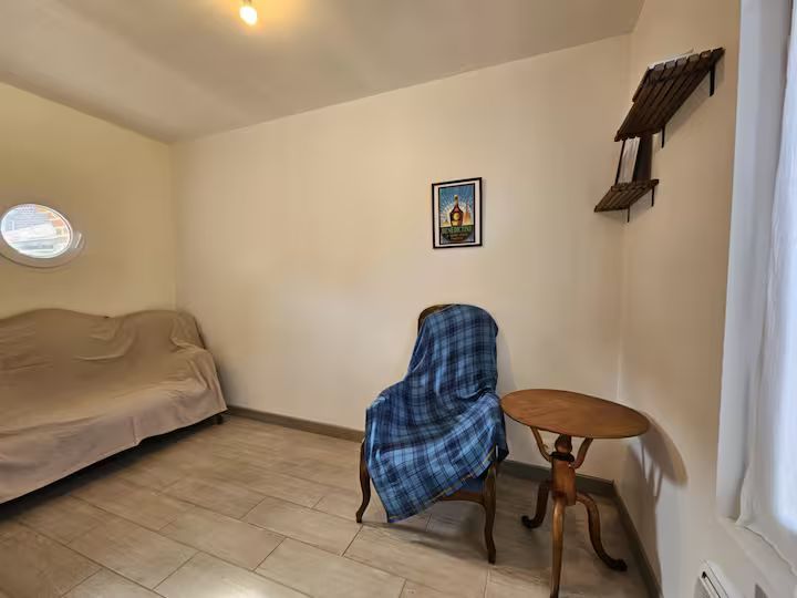 Petite chambre avec un lit, une chaise bleue, une table de chevet ronde, des tableaux et une petite étagère murale.