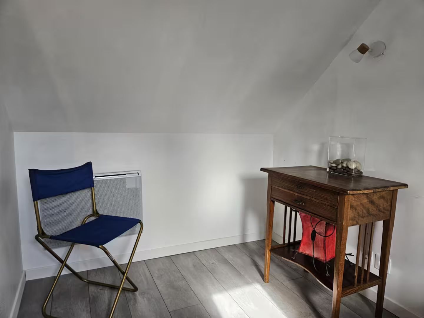 Petite chambre mansardée avec une chaise pliante bleue et une table en bois sous un plafond mansardé.