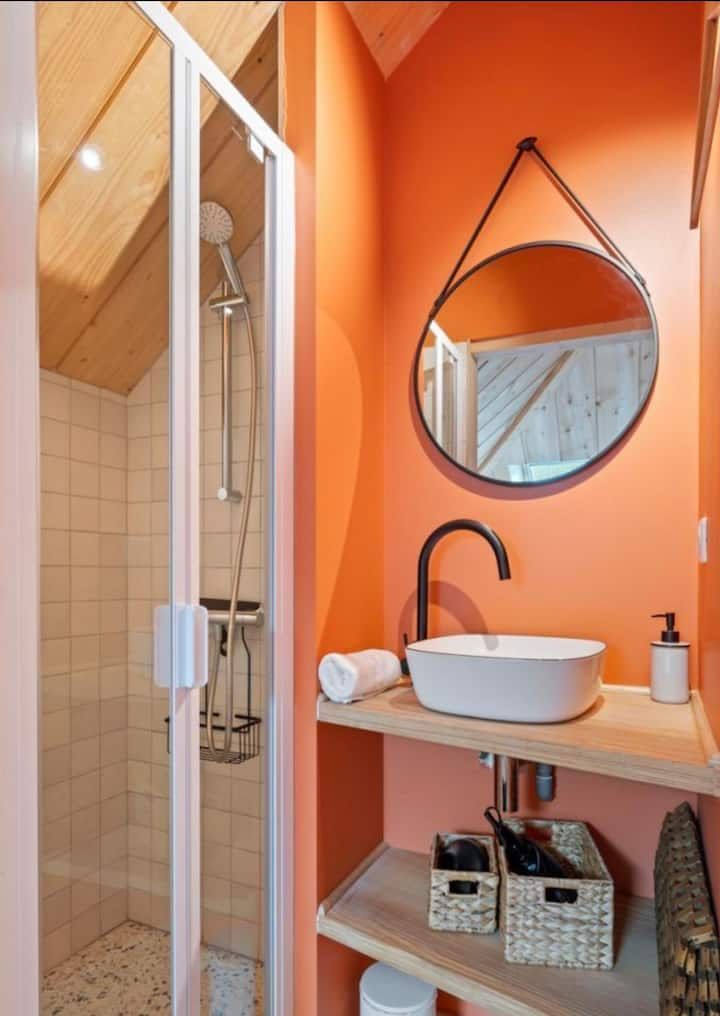 Petite salle de bains orange avec un miroir rond, un lavabo vasque, un robinet noir et une cabine de douche.