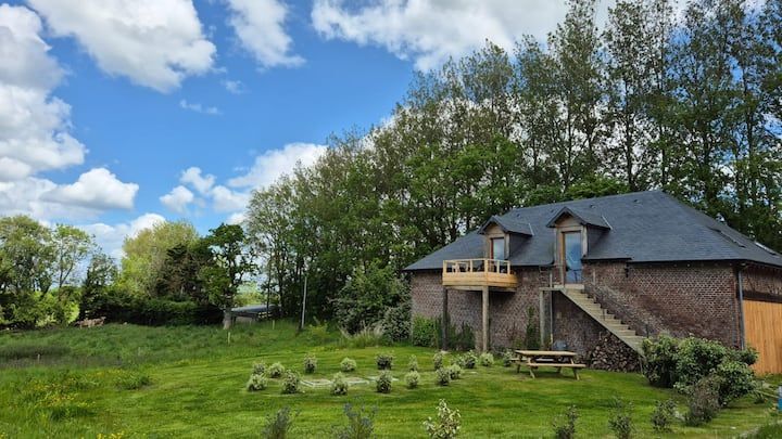 Maison en briques au toit d'ardoise, jouxtant un champ herbeux et des arbres sous un ciel bleu partiellement nuageux.