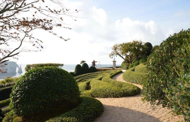 Allée de jardin soignée, bordée de haies et d'arbres taillés, surplombant la mer au coucher du soleil