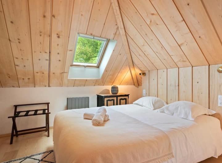 Chambre mansardée confortable avec plafond lambrissé, puits de lumière et grand lit aux draps blancs.