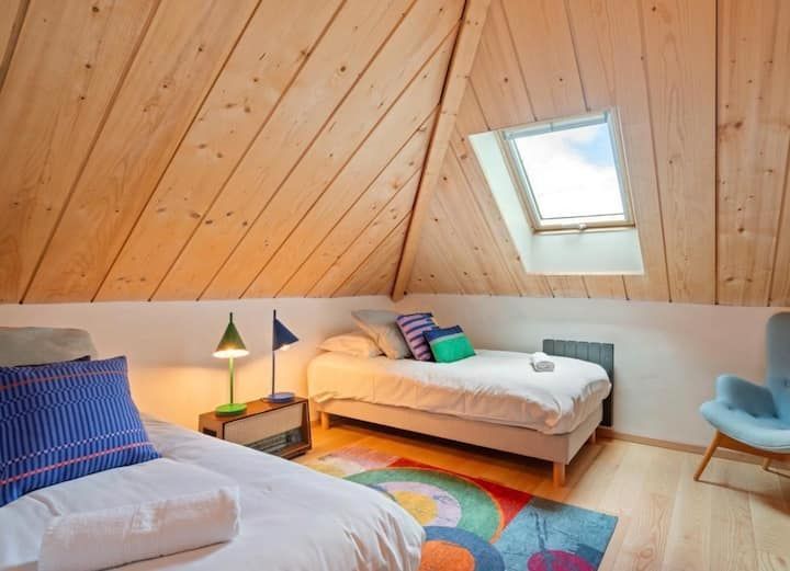 Chambre mansardée confortable avec deux lits, tapis coloré, plafond en bois incliné et puits de lumière