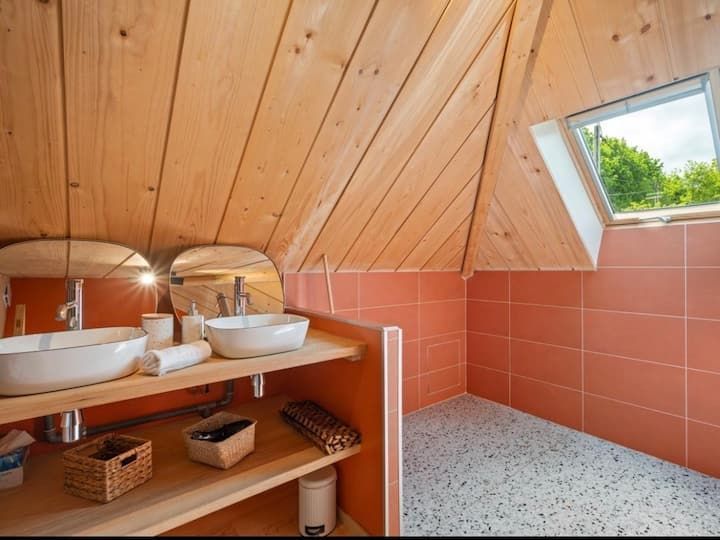 Salle de bains moderne avec plafond en bois incliné, murs en carrelage corail et puits de lumière au-dessus des deux lavabos.