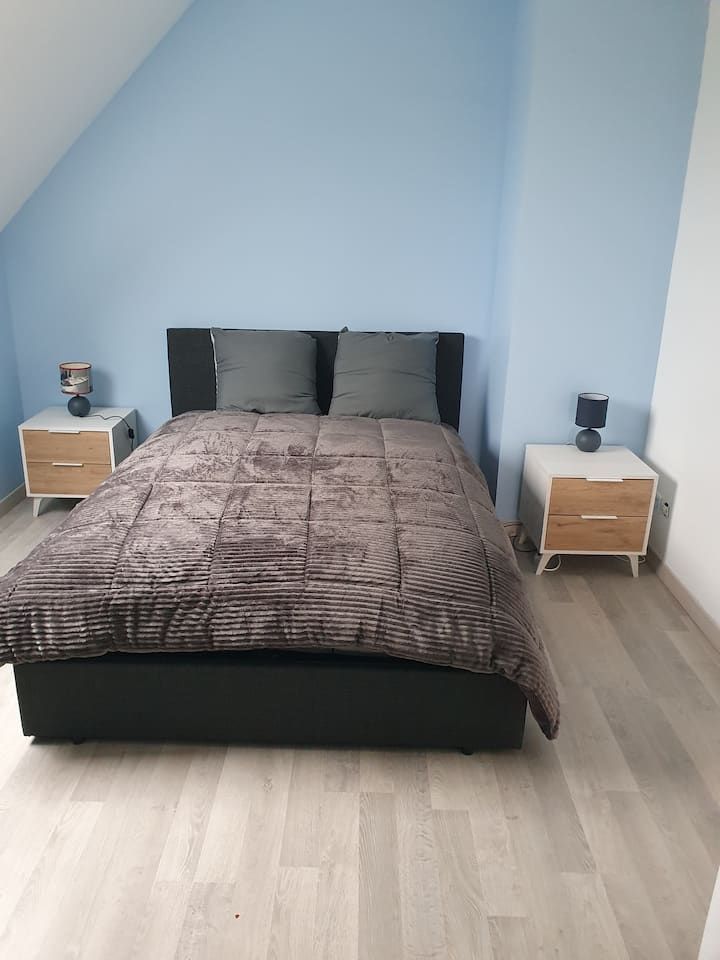 Petite chambre avec un lit, des draps foncés et des tables de chevet assorties sous un plafond bleu en pente.