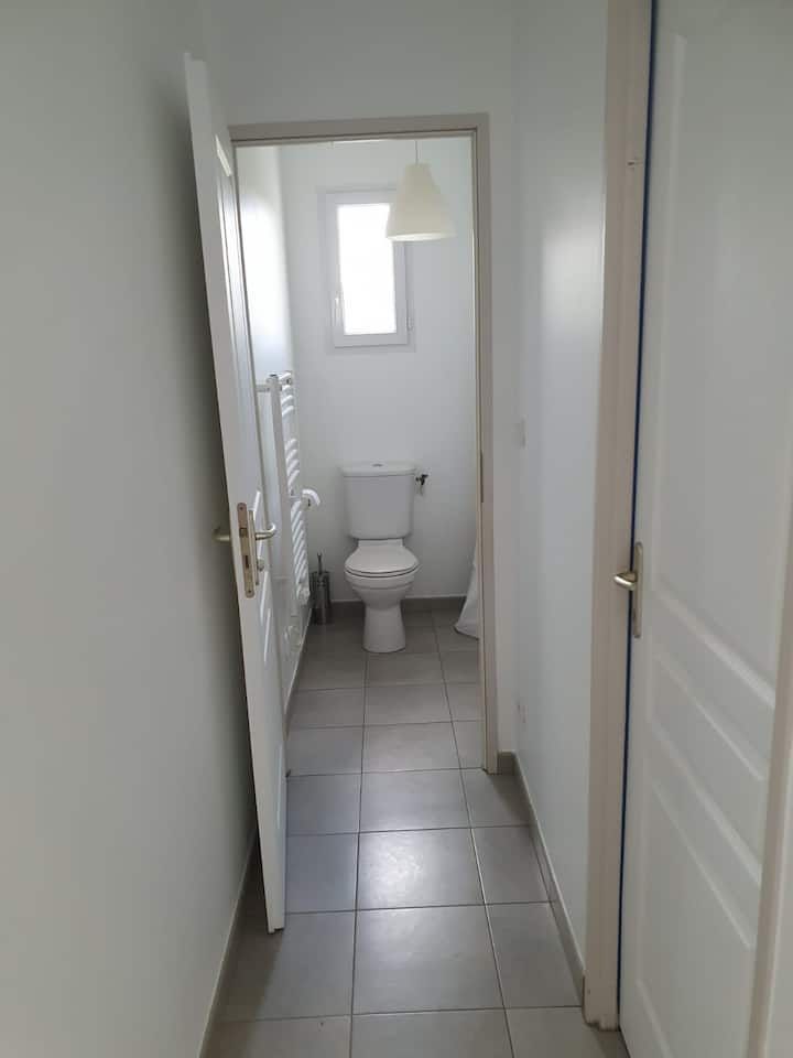 Vue depuis un couloir étroit donnant sur une petite salle de bains avec toilettes, sol carrelé et fenêtre.
