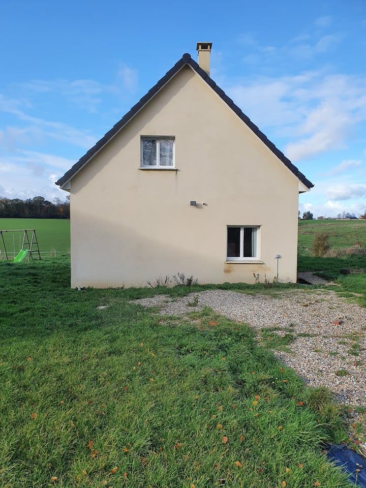 Petite maison beige au toit pentu, située dans un champ herbeux sous un ciel bleu.