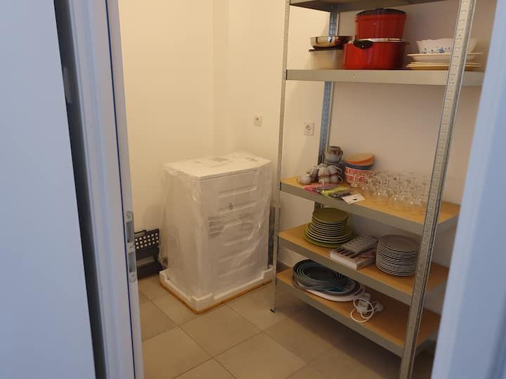 Petit débarras avec des étagères métalliques, des assiettes empilées, des bocaux et un appareil électroménager recouvert, posé au sol.
