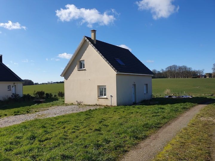 Petite maison couleur crème au toit sombre, située dans un champ ensoleillé et verdoyant sous un ciel bleu.