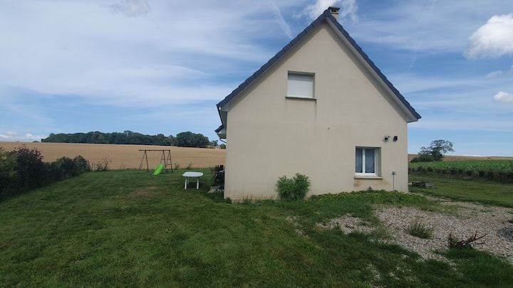 Petite maison beige au toit pentu, située à côté d'un champ herbeux sous un ciel nuageux.