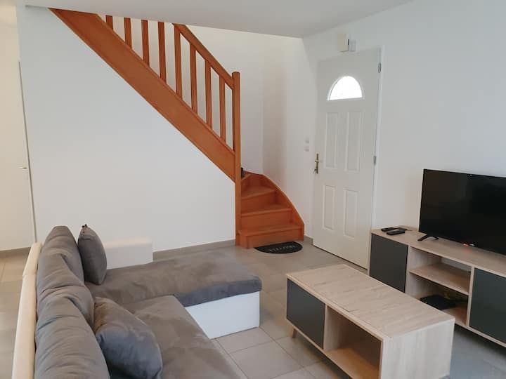 Salon lumineux avec canapé d'angle gris, meuble TV, escalier en bois et porte d'entrée blanche.