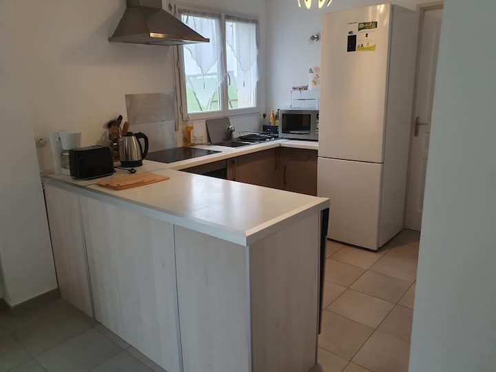 Cuisine blanche compacte avec îlot central, plaque de cuisson, évier et réfrigérateur près d'une fenêtre