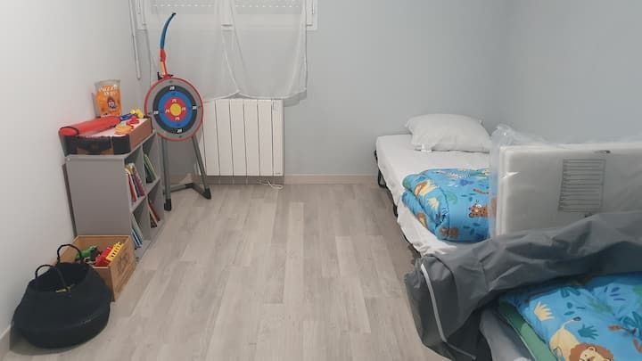 Petite chambre d'enfant avec deux lits jumeaux, une cible pour les jouets et un coin jeux à côté d'un radiateur et d'un rideau.