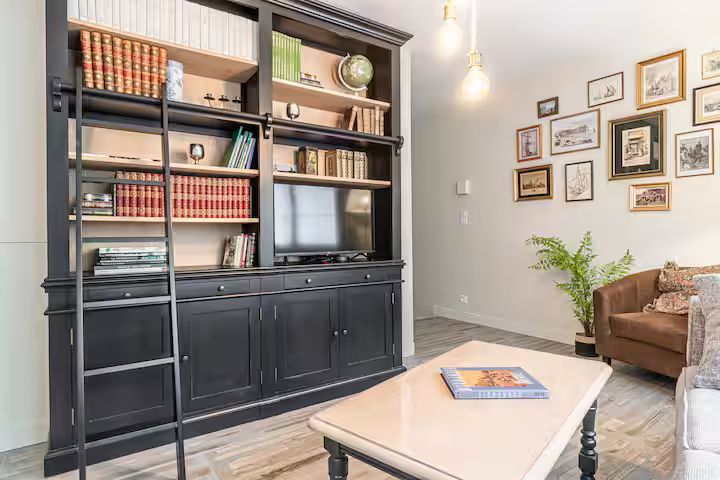 Salon avec une bibliothèque noire, une échelle et une télévision, entourés de tableaux encadrés et d'une table basse.