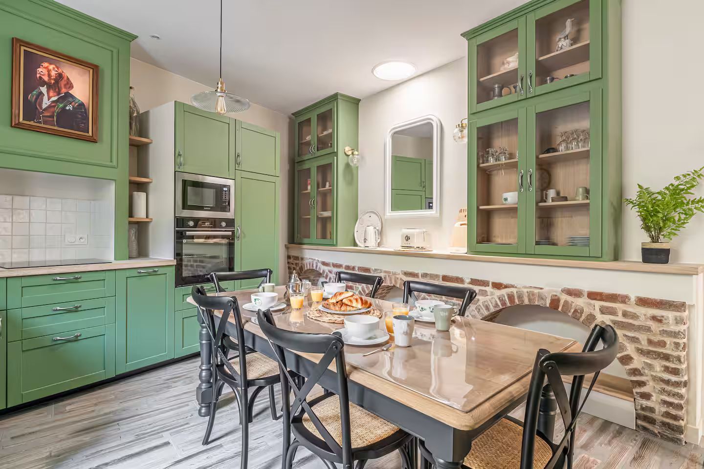 Cuisine verte avec table à manger, éléments en brique et vitrines.