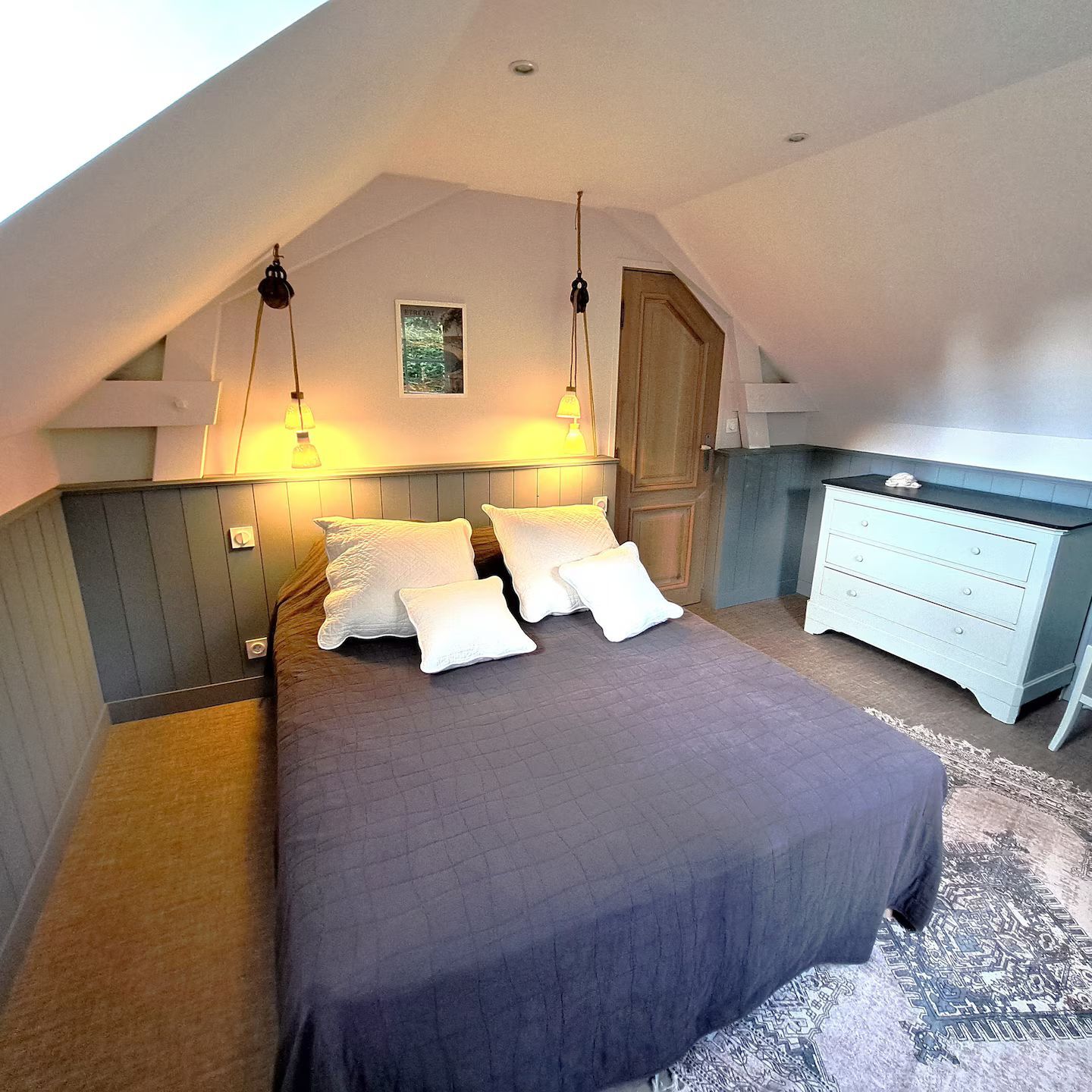 Chambre avec lit, oreillers, lampes et commode. Plafond mansardé et lambris.