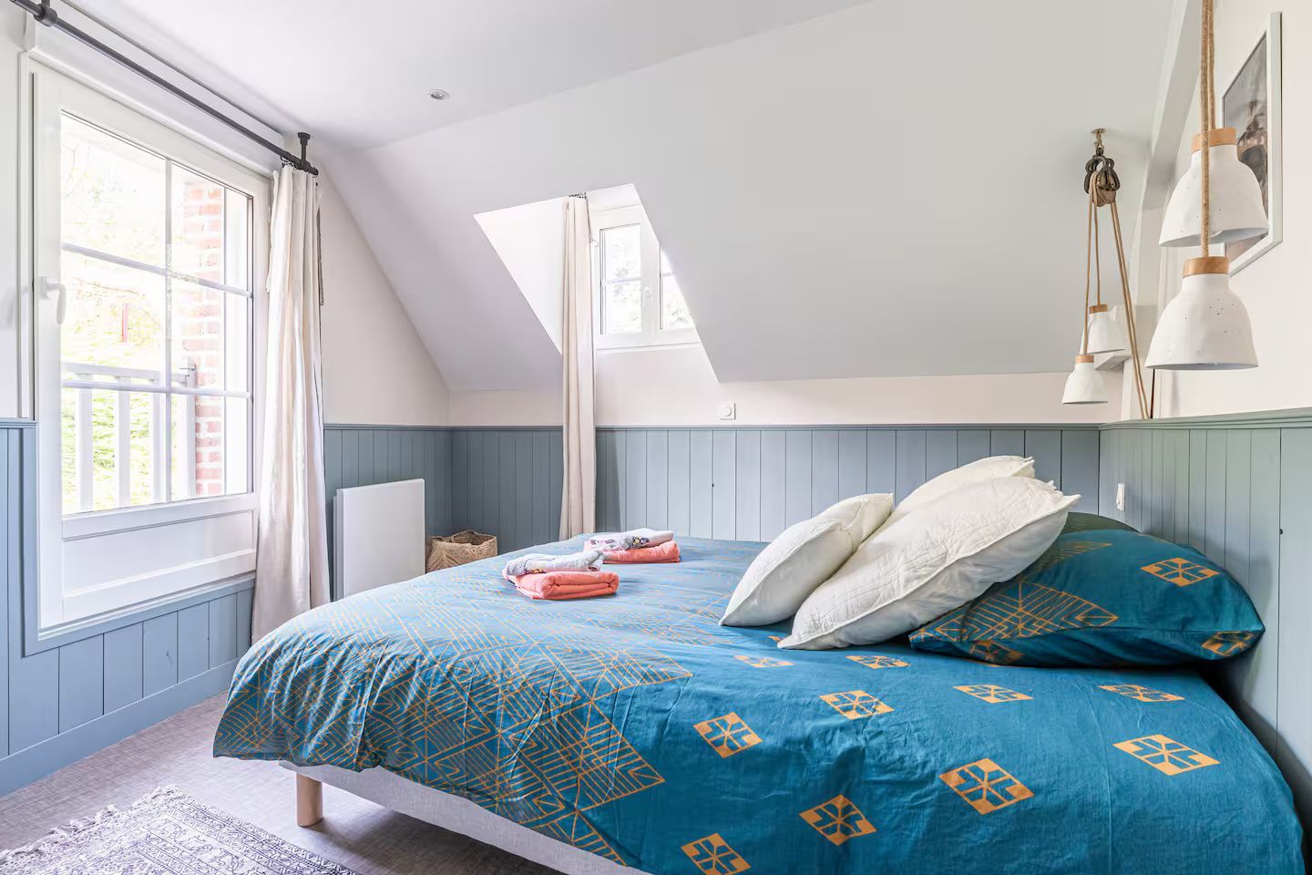 Chambre avec une couette bleue, des oreillers blancs, une fenêtre et des suspensions lumineuses.