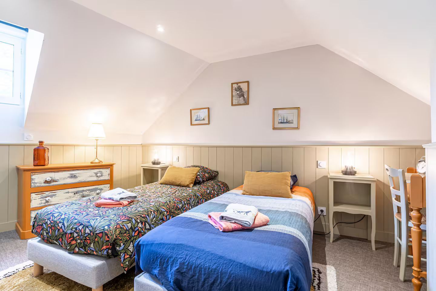Deux lits jumeaux dans une chambre avec plafond mansardé, murs beiges et boiseries ; œuvres d'art encadrées, lampes.