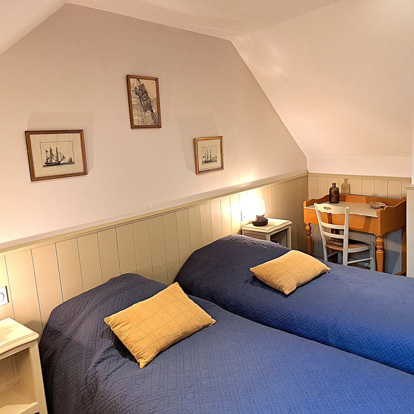 Chambre avec deux lits bleus, des oreillers jaunes, un bureau et des œuvres d'art encadrées.