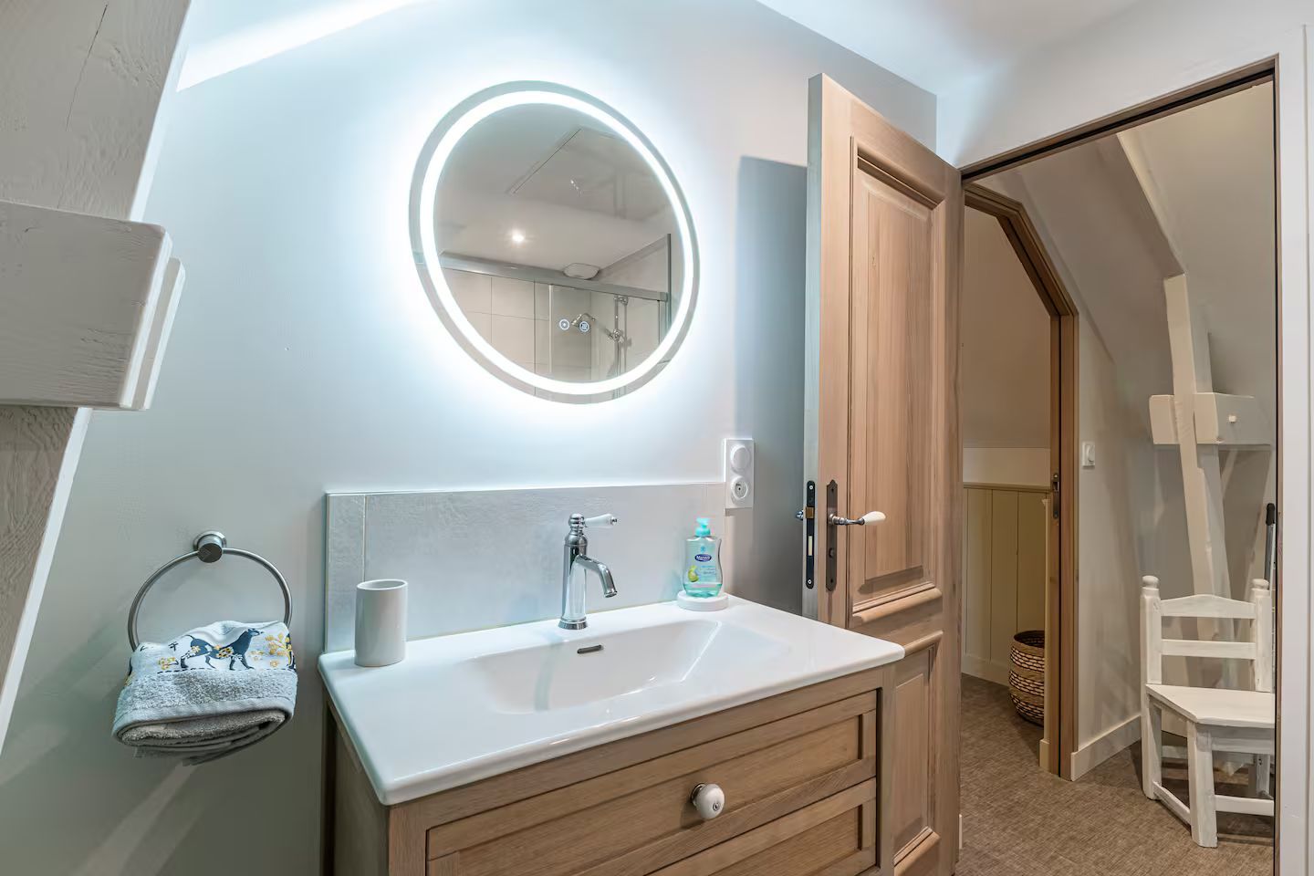 Salle de bains avec lavabo blanc, miroir éclairé, armoire en bois et porte ouverte donnant sur une petite pièce.