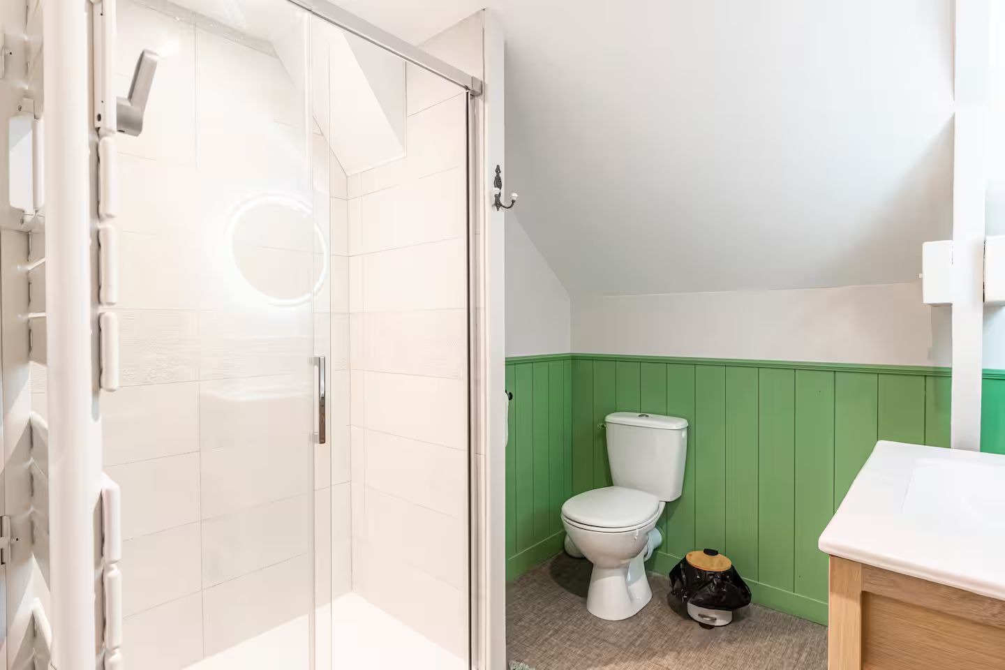 Salle de bains avec douche en verre, toilettes contre des lambris verts et plafond blanc en pente.