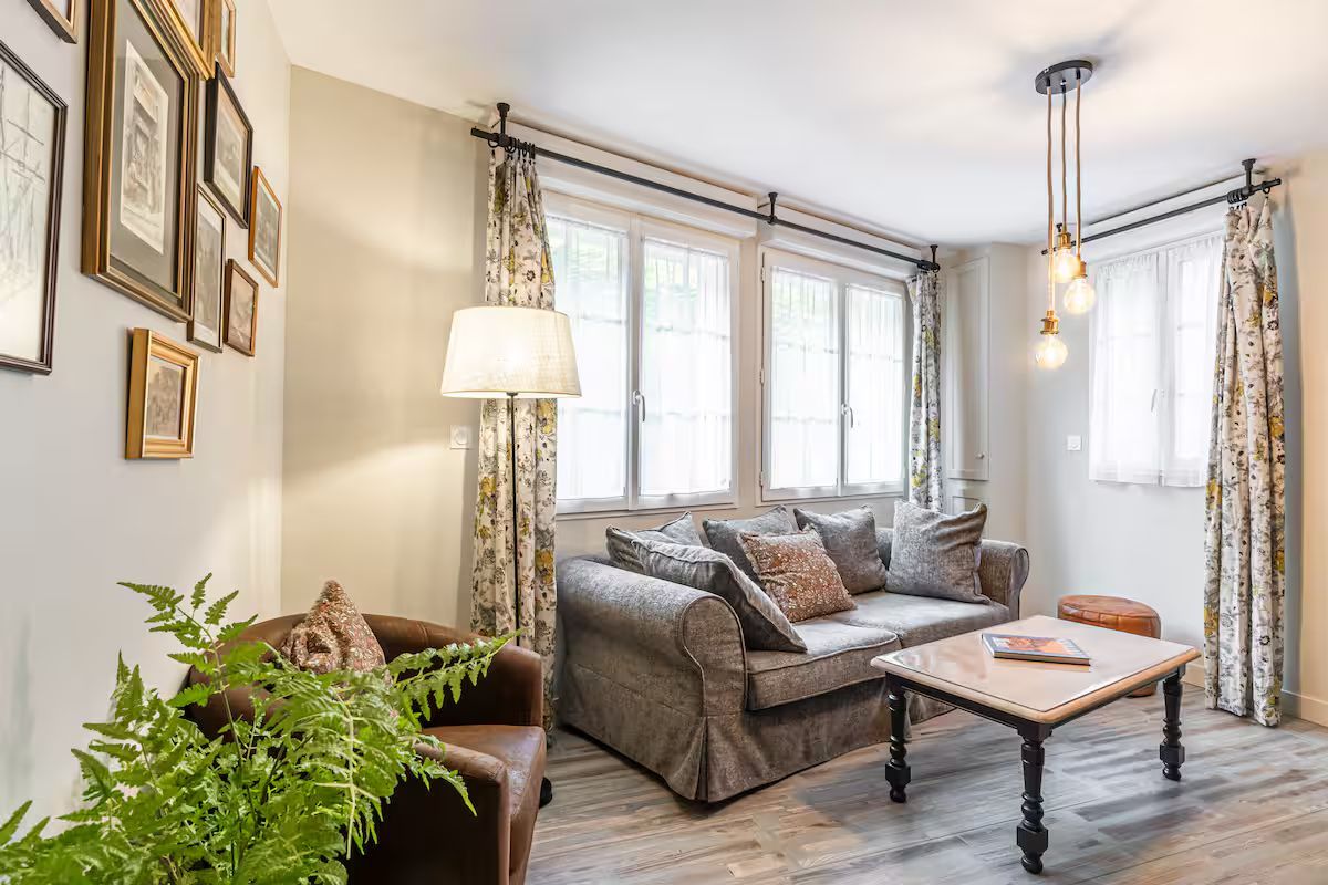 Salon cosy avec canapé gris, fauteuil marron, rideaux à fleurs, mur de cadres et table basse.