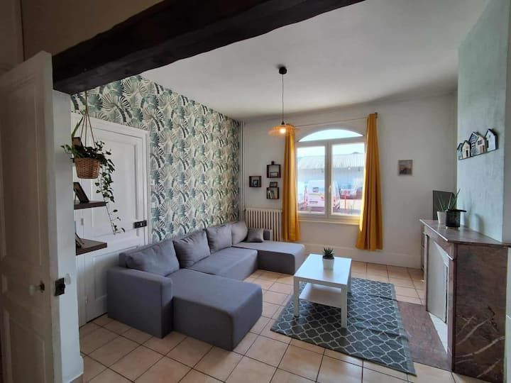 Salon avec canapé d'angle gris, papier peint à motifs, table basse, cheminée et fenêtre avec rideaux jaunes.