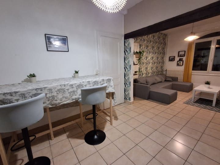 Salon moderne avec sol carrelé, canapé gris, petite table basse et deux tabourets de bar près d'un comptoir.