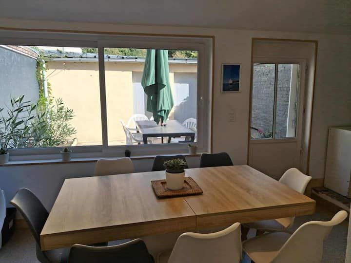 Salle à manger avec table en bois, chaises noires et grande fenêtre donnant sur une terrasse avec parasol.